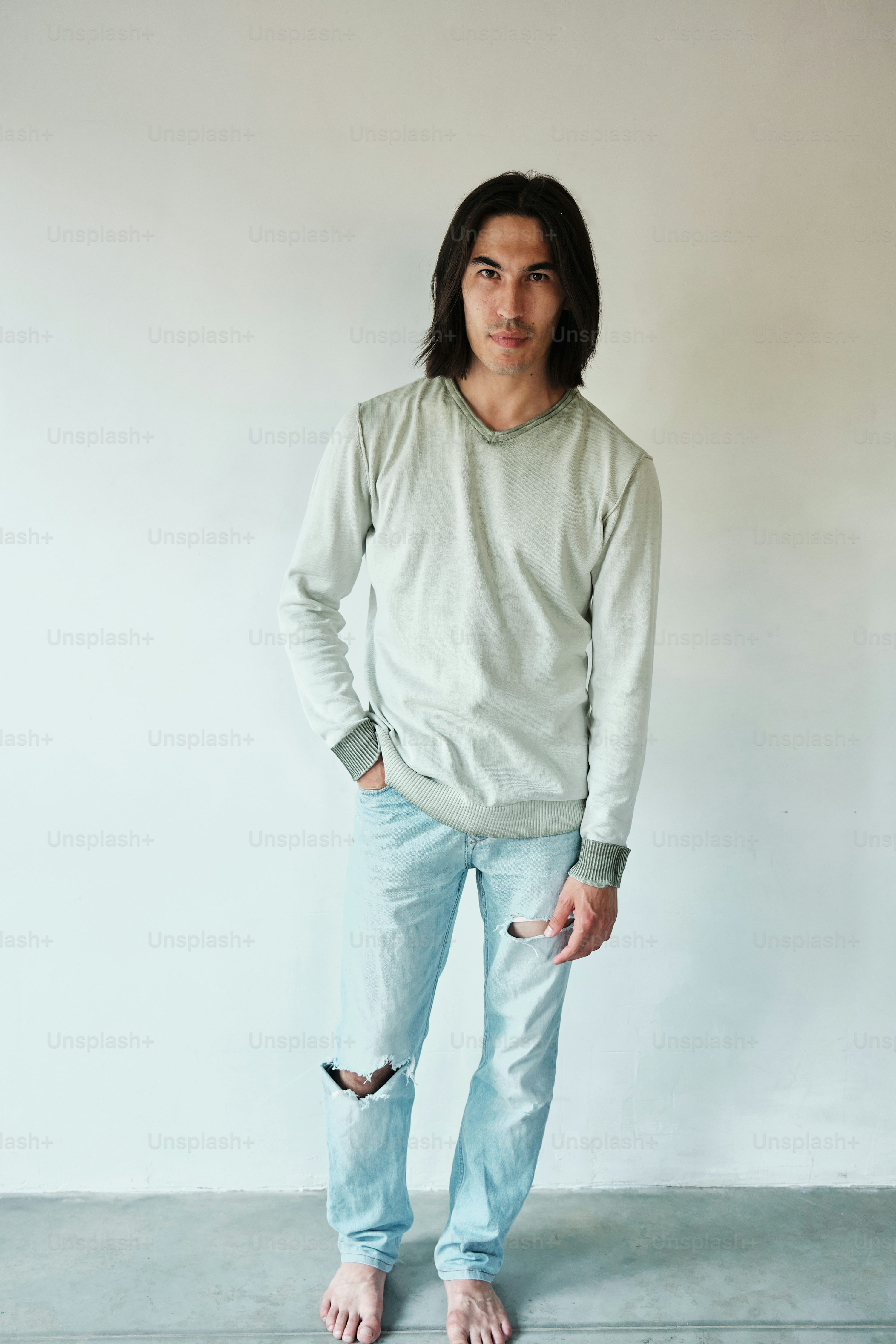 a young asian man with long brown hair stands barefoot against a white wall. he's wearing a light grey longsleeved sweater and light blue jeans with rips at the knees. he has one hand in his pocket and is looking directly at the camera. the overall style is casual and relaxed.