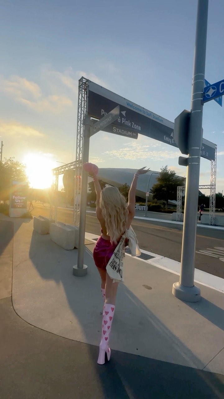 a woman in pink boots and a dress stands near sofi stadium at sunset. she is holding her hands up in the air, and she is carrying a bag. there is a sign in the background.