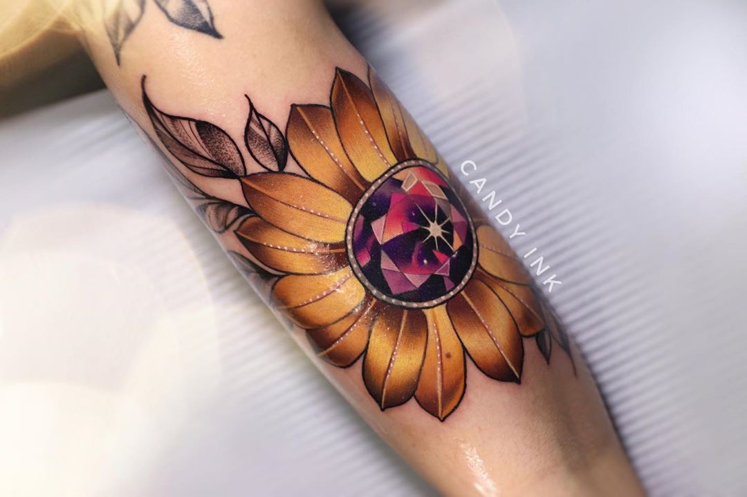 a closeup shot of a freshly done tattoo on the back of someone's arm. the tattoo features a sunflower with a large, multifaceted purple and pink jewel in the center. the petals of the sunflower are a bright golden yellow, and there are a few smaller leaves in shades of green and gray tattooed around the flower. the tattoo artist's watermark, "candy ink," is visible in stylized letters along the right side of the tattoo. the photo is taken indoors in a tattoo parlor with artificial lighting.