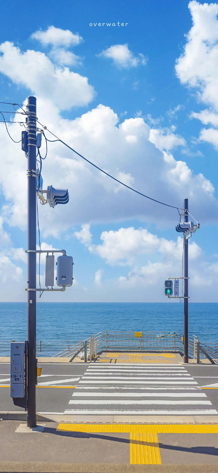 the image shows a crosswalk on a street leading towards the ocean. there are two utility poles with crosswalk signals on either side of the street. the crosswalk is marked with white stripes and there is tactile paving in bright yellow on the sidewalk leading up to it. a traffic light shows a green walking person symbol, indicating it is safe to cross. the ocean stretches out in the background with a blue sky and puffy white clouds above. the word "overwater" is written at the very top center of the frame.