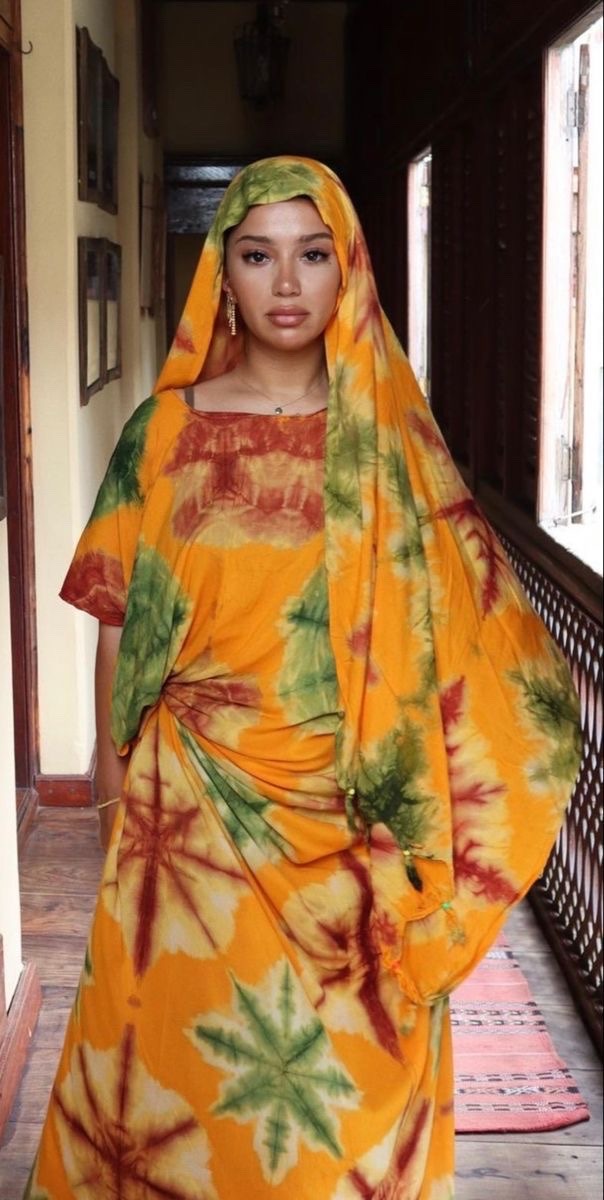 a woman stands in a hallway wearing a colorful tiedye dress and headscarf. the dress is yellow, orange, green, and red. she is also wearing earrings and a necklace. the hallway is decorated with brown wood and there is a rug on the floor. she is looking directly at the camera.