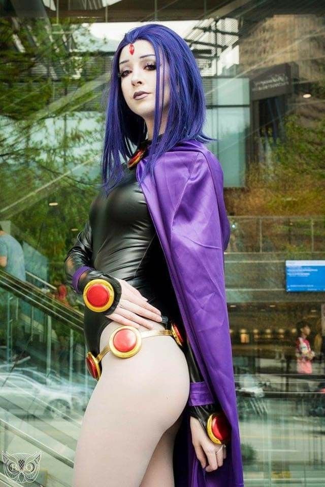 the image features a woman dressed as raven, a dc comics superheroine. she has purple hair and a red gemstone on her forehead, and is wearing a black bodysuit with red and gold accessories. she also has a purple cape draped over her shoulders. the background appears to be an indoor location, possibly a convention or event. the woman is looking directly at the camera.