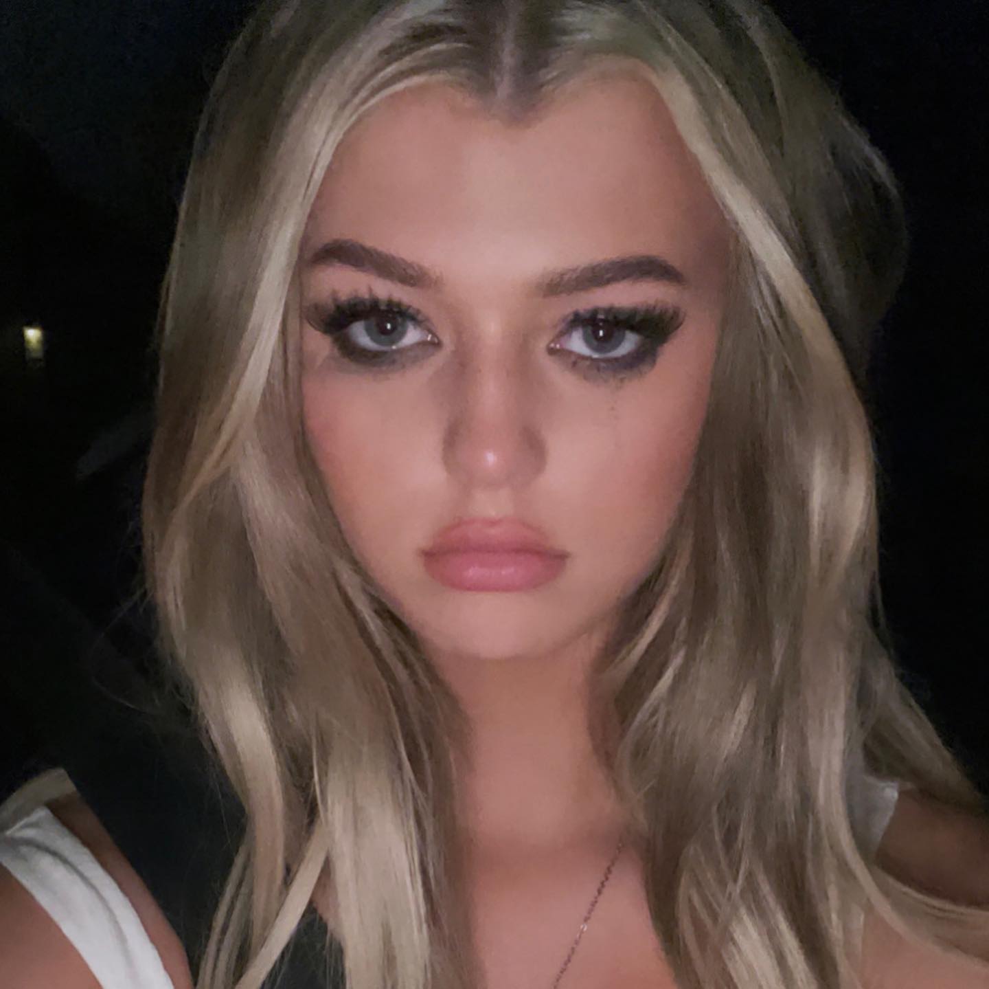 a closeup selfie of a woman with blonde hair, dark eye makeup, and fair skin. she is wearing a necklace and has gray eyes. the background is blurred. she has thick eyeliner on her eyes and long false eyelashes.