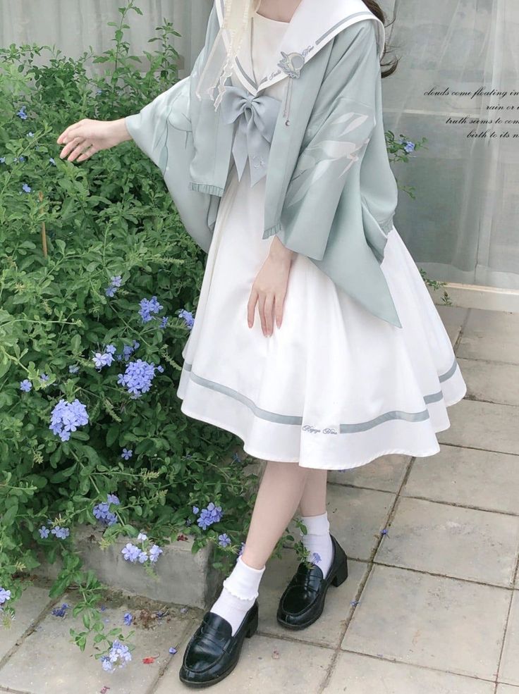 a woman is standing outdoors in front of a wall and some plants. she is wearing a white dress with a mint green jacket over top. the jacket has a sailor collar and a large bow on the front. she is also wearing white socks and black loafers. the overall style of her outfit is lolita fashion, which is a japanese street style that is characterized by its dolllike aesthetic.