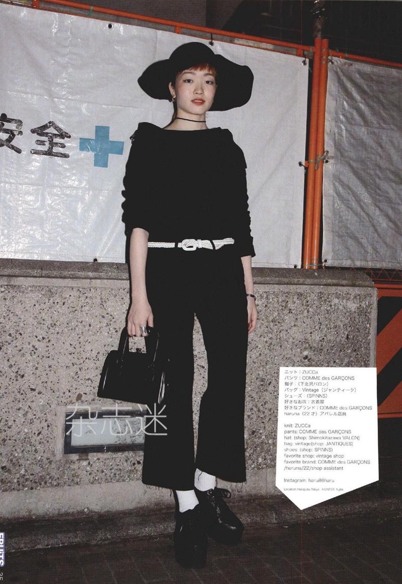 a young woman is posing on a sidewalk in front of a construction site. she is wearing a black hat, black top, black wideleg pants, white socks, black platform shoes, and carrying a black handbag. she has short black hair and is looking at the camera. the image is welllit with natural daylight.  the overall style is a mix of vintage and modern japanese street fashion with an emphasis on black and white.  her outfit incorporates pieces from the japanese fashion label comme des garcons.