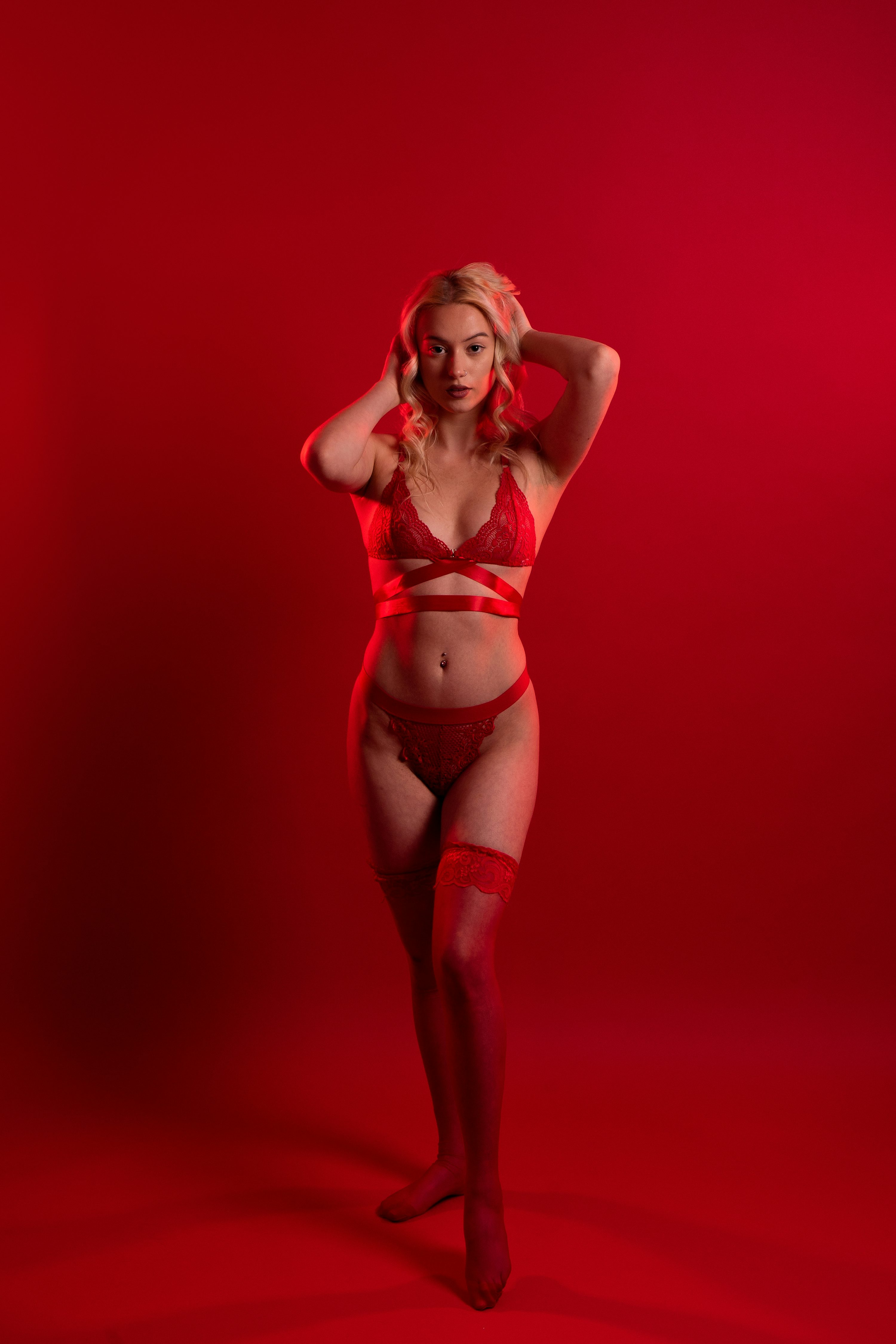 the image features a blonde woman standing against a solid red background. she is wearing a red lace lingerie set consisting of a bra and panties, along with matching red stockings.  her hands are placed gently behind her head. the overall lighting is red, emphasizing the color of her clothing and creating a sultry, romantic atmosphere. the image is clearly a studio portrait, showcasing the model and her attire in a glamorous, sensual way.