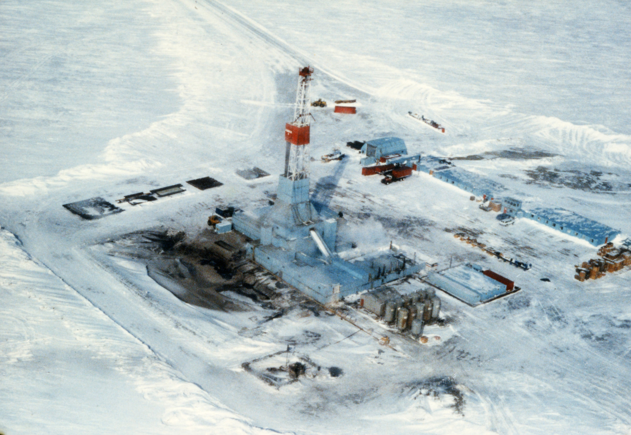 an aerial view of an oil rig drilling operation in a snowy, icy arctic landscape. the rig is visible in the center of the image, with various support structures and equipment scattered around it. the ground is covered in snow and ice, and the surrounding landscape is desolate and cold. the rig's tower rises high above the ground, signifying the challenging and harsh environment in which it operates. this image captures the vastness and remoteness of the arctic, as well as the industrial operations that take place in this region.