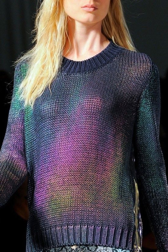 a blonde woman model is wearing a dark, iridescent sweater on a runway. she is looking ahead with a serious expression. the sweater is loose fitting and the knit fabric shimmers with a purple, blue, and green effect.