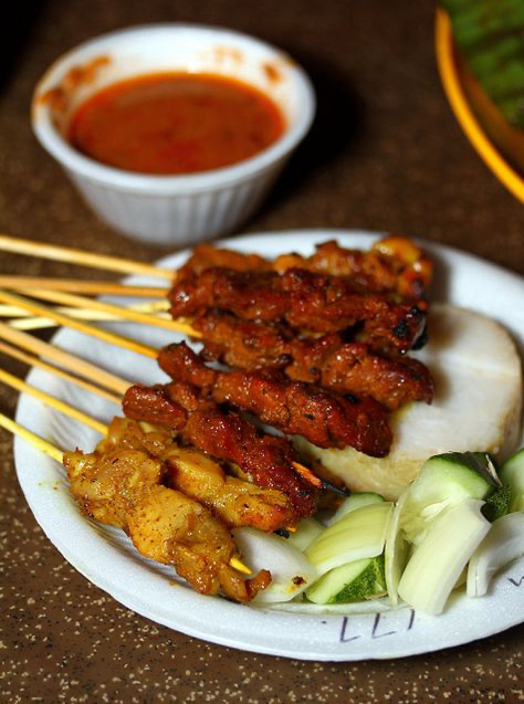 a closeup shot of a plate of satay, a popular southeast asian dish. the plate contains several skewers of grilled chicken and pork satay, accompanied by ketupat (rice cakes) and slices of cucumber and onion. a small bowl of rich, reddishbrown peanut sauce is visible in the background, along with a blurry yellow plate in the upperright corner. the satay looks appetizing, with a slightly charred exterior, suggesting delicious grilling. the overall setting suggests a street food vendor or casual outdoor eatery. the photo is welllit and showcases the textures and colors of the food beautifully.