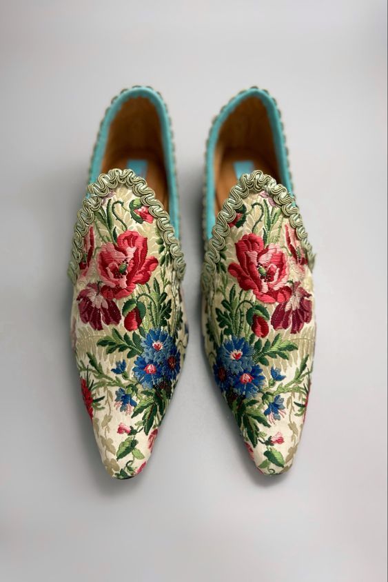a pair of floral embroidered pointedtoe loafers with turquoise trim are seen from a topdown perspective on a light gray surface. the shoes are intricately embroidered with a pattern featuring red roses and blue flowers, set against a creamcolored background. the turquoise trim lines the opening of the shoe, adding a pop of color.
