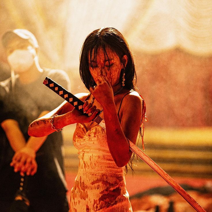 a closeup shot of a young asian woman, covered in blood, clutching a katana.  her white dress is heavily stained with blood. she appears to be in distress, possibly injured or wounded. a blurred figure of a masked man is visible in the background, suggesting a scene of violence or a fight. the overall tone is dark, dramatic, and cinematic, resembling a still from an action thriller or horror film. the lighting is intense, emphasizing the blood and the woman's emotional state.