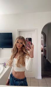 a selfie of loren gray in a bathroom. she is holding her phone up to the mirror. she is wearing a white crop top and jeans. there is a tv on the wall behind her. there is a sink to her left. there is a door behind her and an arched doorway to the right.