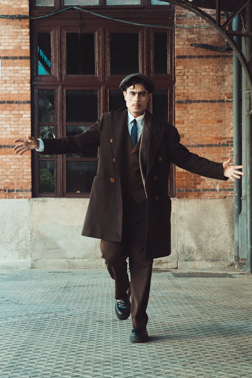 a man in a brown suit and overcoat, wearing a hat and necktie, is standing indoors in front of a window and a brick wall. he has his arms outstretched.