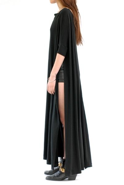 a woman stands in profile, wearing a long, black dress with a side slit that reveals her leg. she has a bracelet on her wrist and is wearing black ankle boots.