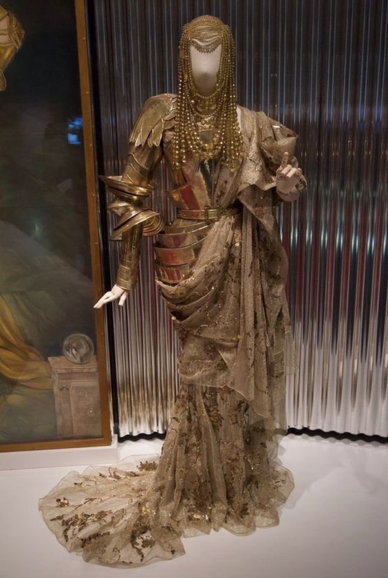 a mannequin is wearing an extravagant gold and metallic gown, covered in glitter and gold. the gown is flowing and has a long train. the mannequin is also wearing jewelry and a gold crown. the mannequin is on display in a museum or gallery.