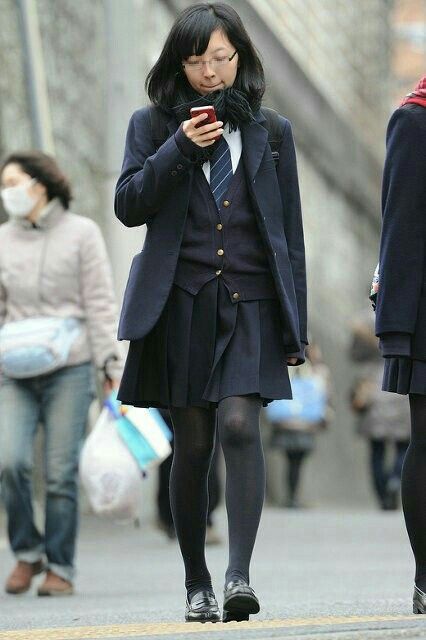 a young woman in a japanese school uniform is walking down a city sidewalk while looking at her smartphone. she is wearing a navy blue blazer, a pleated skirt, black tights, and black loafers. her hair is dark and straight, and she is wearing glasses. the background is slightly blurred, with other pedestrians walking in the opposite direction. the overall color palette is muted, with shades of gray, blue, and black dominating the scene.