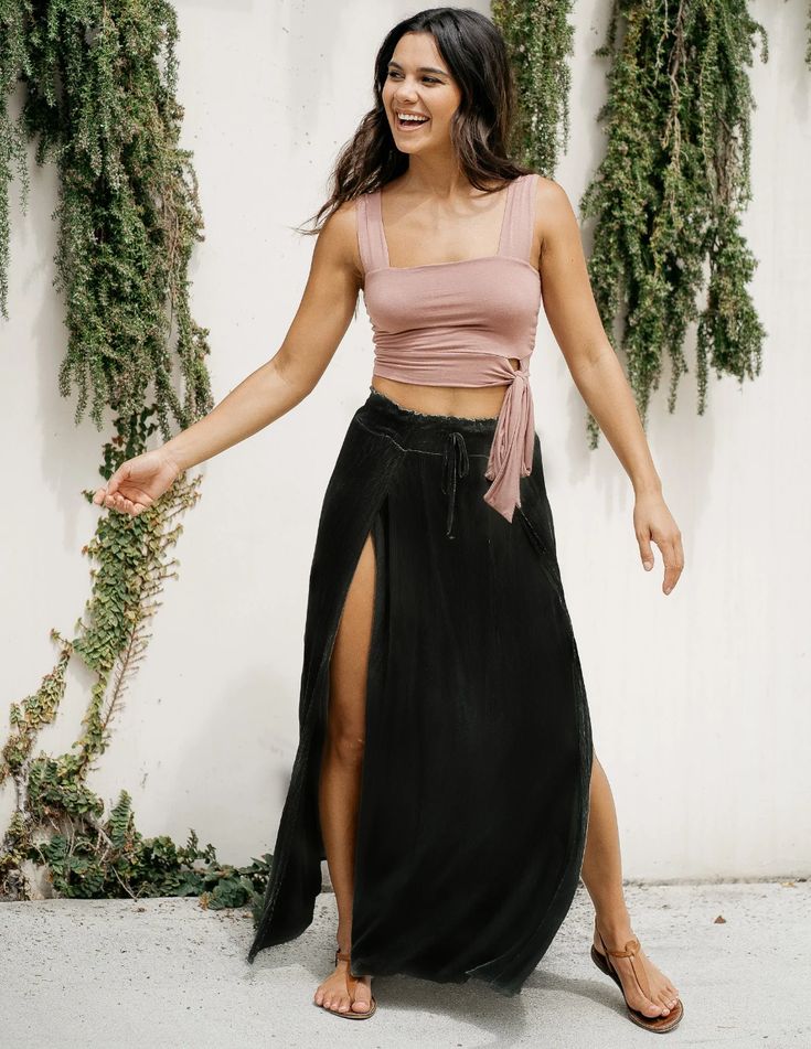 a woman wearing a dusty pink crop top and a long black skirt with two thighhigh slits, is standing outside in front of a white wall with green vines. she is smiling and looking to the side. she is also wearing brown sandals. the image is welllit with natural sunlight.