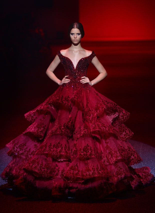 a young woman in a red gown is posing on a runway. the gown is a beautiful and elegant design, with lace, ruffles, and sparkles. the model is confident and graceful, and her pose is striking. the background is a red carpet, which adds to the glamour of the occasion. this photo is a perfect example of haute couture fashion.