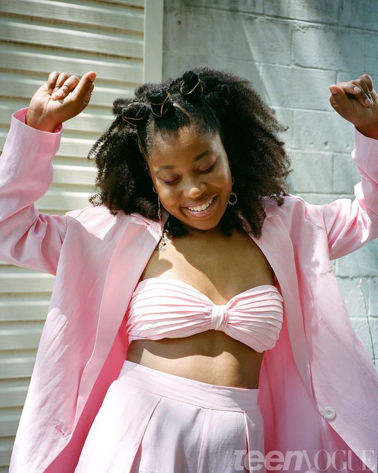 a young black woman with big natural hair styled with two space buns is joyfully dancing in the sun. she is wearing a pink threepiece outfit that includes a blazer, bandeau top, and pants.  the background is a white building exterior with a brick wall.