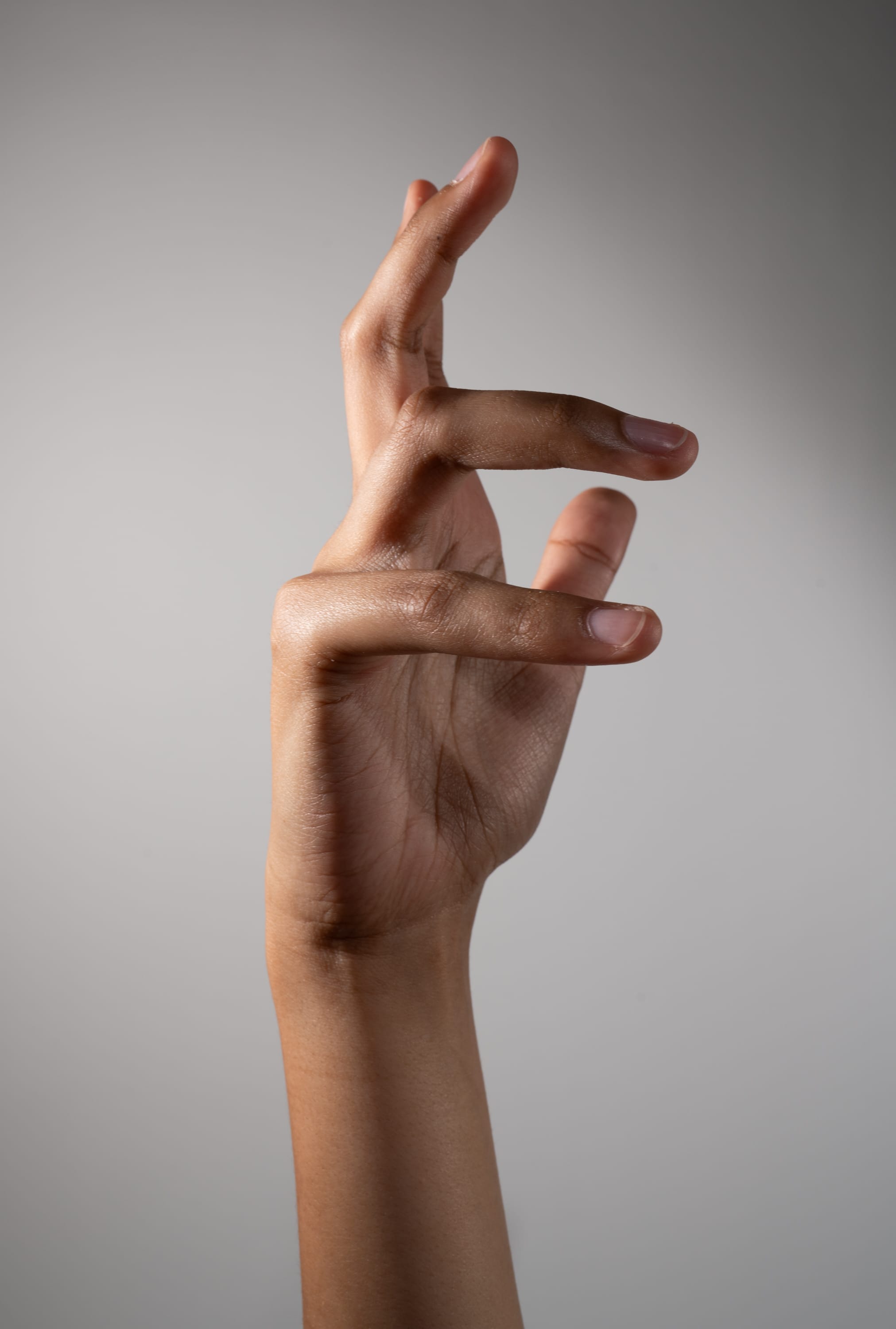 a single hand of a woman with brown skin is extended upwards, fingers curled in a graceful gesture against a muted grey background.  the hand is elegantly lit with soft studio lighting.