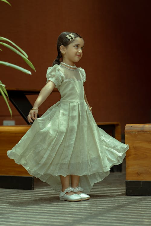 a young girl is wearing a light green dress with short puffed sleeves and a pearl necklace. she has a hair accessory in her hair and is posing with a slight smile. the dress is full and flows out around her, and she is wearing white shoes. the background is a combination of brown and natural elements such as green plants and wood.