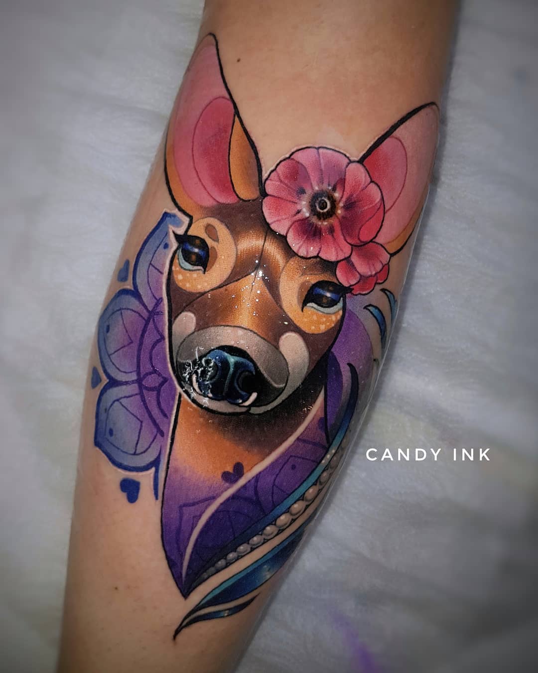a tattoo of a deer's head with flowers on its head, set against a backdrop of a mandala pattern in shades of purple. the deer is rendered in brown and pink hues, with glitter accents. the overall style is whimsical and illustrative.