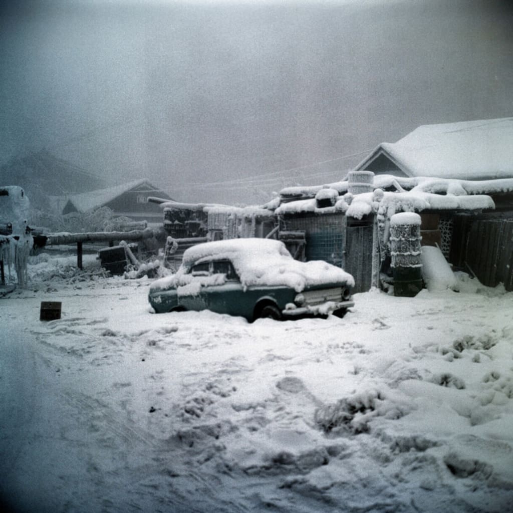 a vintage car is parked in a snowy landscape, with a house in the background and snow covering everything. the car is covered in snow, and the ground is completely white. the sky is overcast and grey. the image evokes a sense of wintertime coldness and isolation.