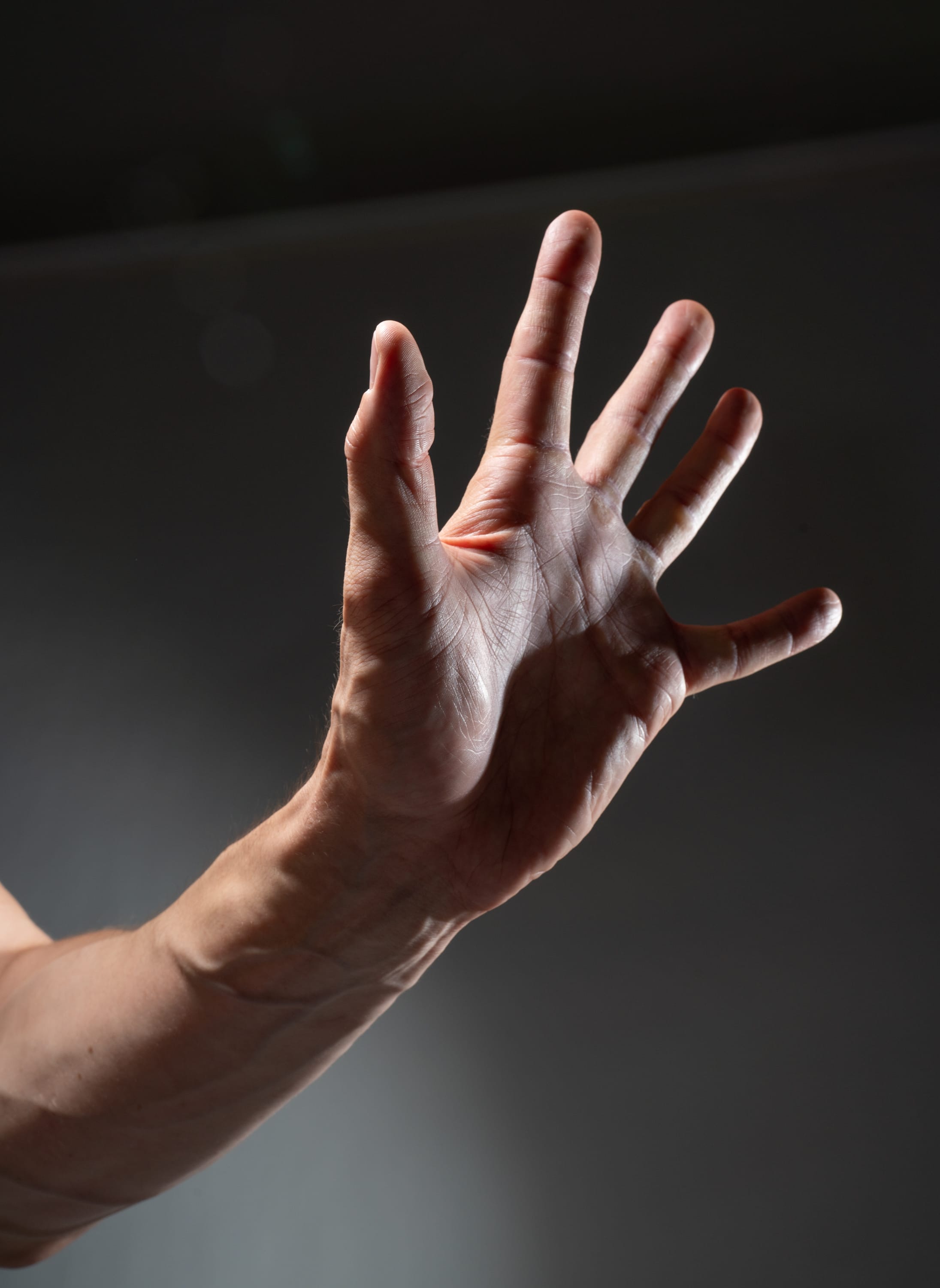 the image shows a man's left hand extended outward, illuminated by a beam of natural light against a dark background. the light accentuates the details of the hand's skin texture, veins, and fingers. the man's arm is visible, suggesting a pose of reaching out or signaling.