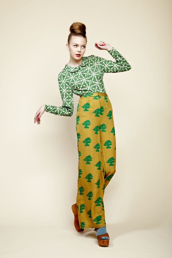 a fulllength studio shot of a fashionable young woman posing in retro clothing. she is wearing a green geometric patterned blouse with a peter pan collar, paired with highwaisted wideleg trousers with a repeating pattern of green stylized trees on a mustard yellow background. her hair is styled in a high bun, and she is wearing brown platform shoes with light blue socks. the model's pose and the clothing's style evoke a 1960s or 1970s aesthetic. the background is a simple, neutral beige.