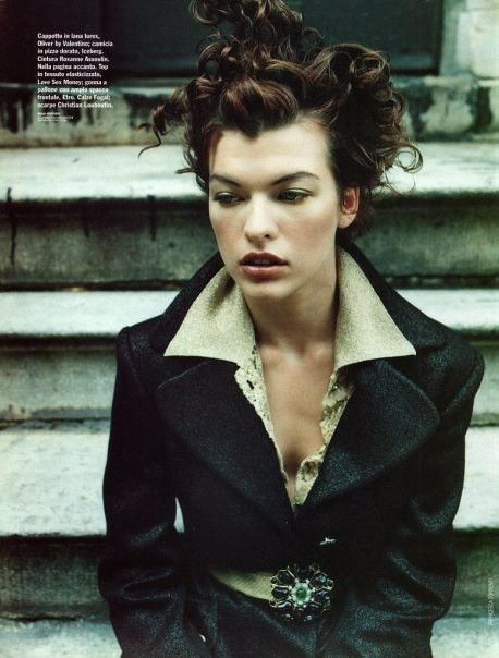 a portrait of milla jovovich wearing a black coat while posing on stairs. the coat is belted with a decorative brooch. her hair is styled in loose, curly waves. the overall style suggests a vintage fashion aesthetic.