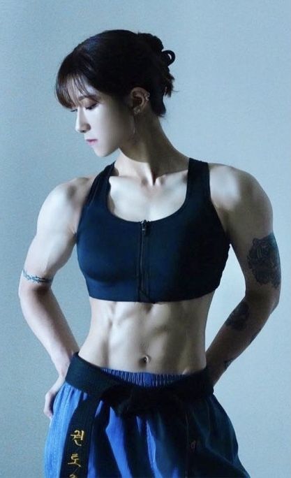 a muscular woman in a black sports bra and blue martial arts uniform stands with her arms crossed in front of her. she has a sixpack and is looking down at the ground. she is wearing a black belt.  her muscles are defined and she is in great shape. the woman has a tattoo on her left arm.