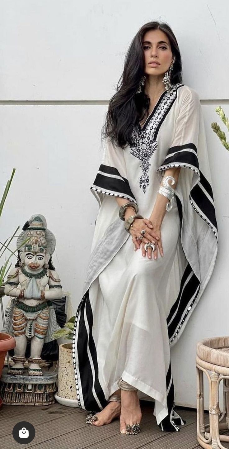 a woman is sitting on the floor, wearing a long white kaftan with black stripes and embroidery. the kaftan has long sleeves and a loose fit. the woman is wearing several pieces of jewelry, including earrings, bracelets, and rings. her hair is long and black, and she is wearing sandals. she is sitting in front of a statue of a hindu god. the background is a white wall with some plants.