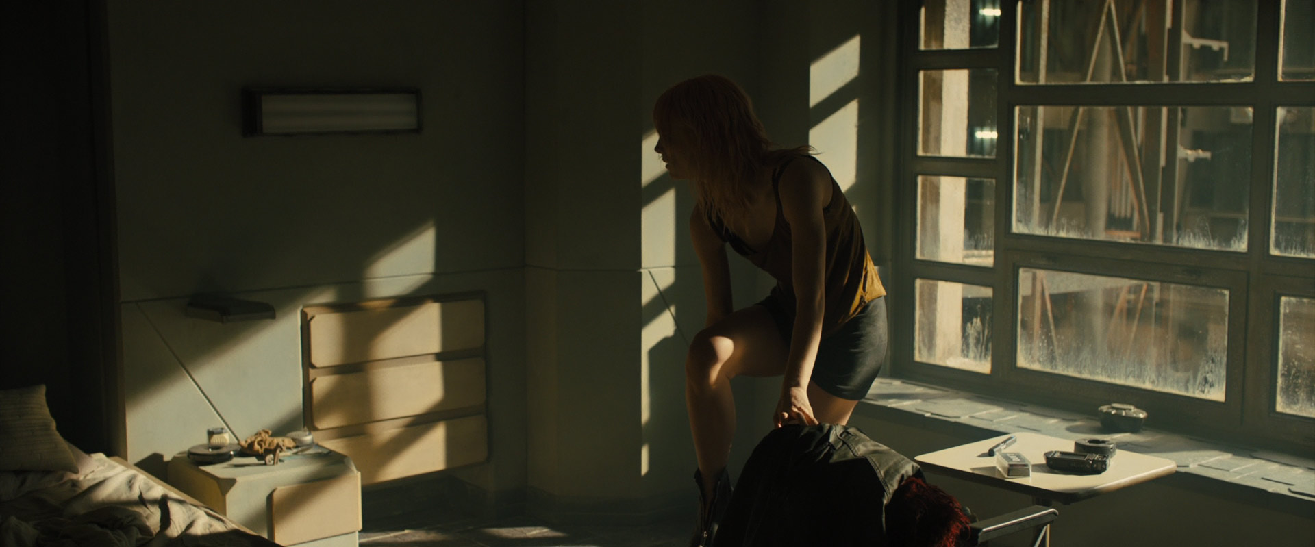 a blonde woman is standing in a dark room with a window. the woman is wearing a yellow tank top and black shorts. she is looking out the window. the room is dusty and dirty and there is a bed to the left with a side table. there are shadows across the room from the sunlight coming in the window.