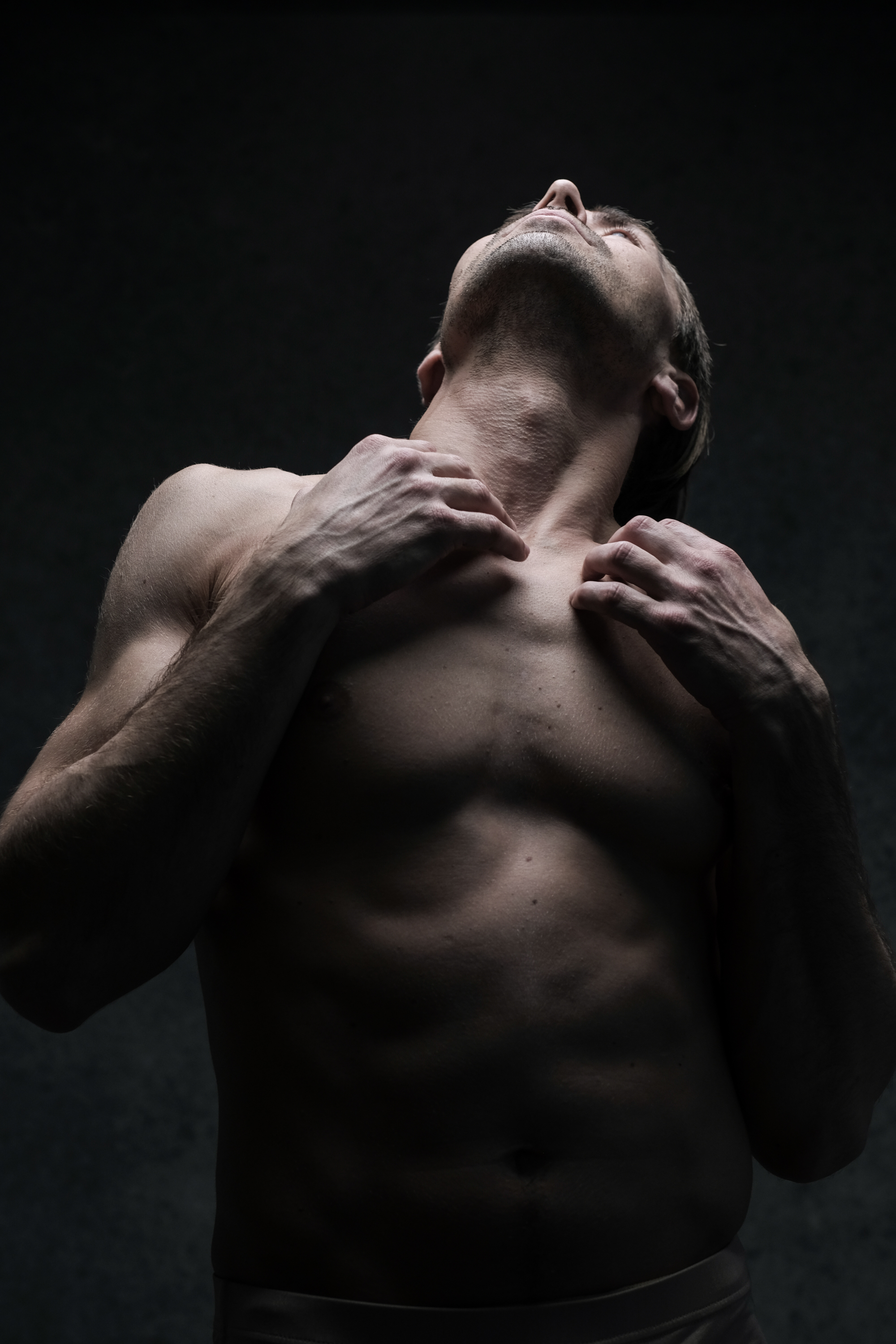 a shirtless man stands with his head tilted back, eyes closed, and hands on his chest, lit dramatically against a dark background. the lighting emphasizes the contours of his muscular physique. his pose suggests a sense of vulnerability and emotional intensity.