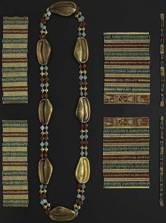 a flatlay image of a collection of ancient egyptian jewelry laid out on a dark gray surface. there are four bracelets on the left side, a long beaded necklace in the center, and three bracelets on the right side. the jewelry is made of gold, carnelian, turquoise, and lapis lazuli.