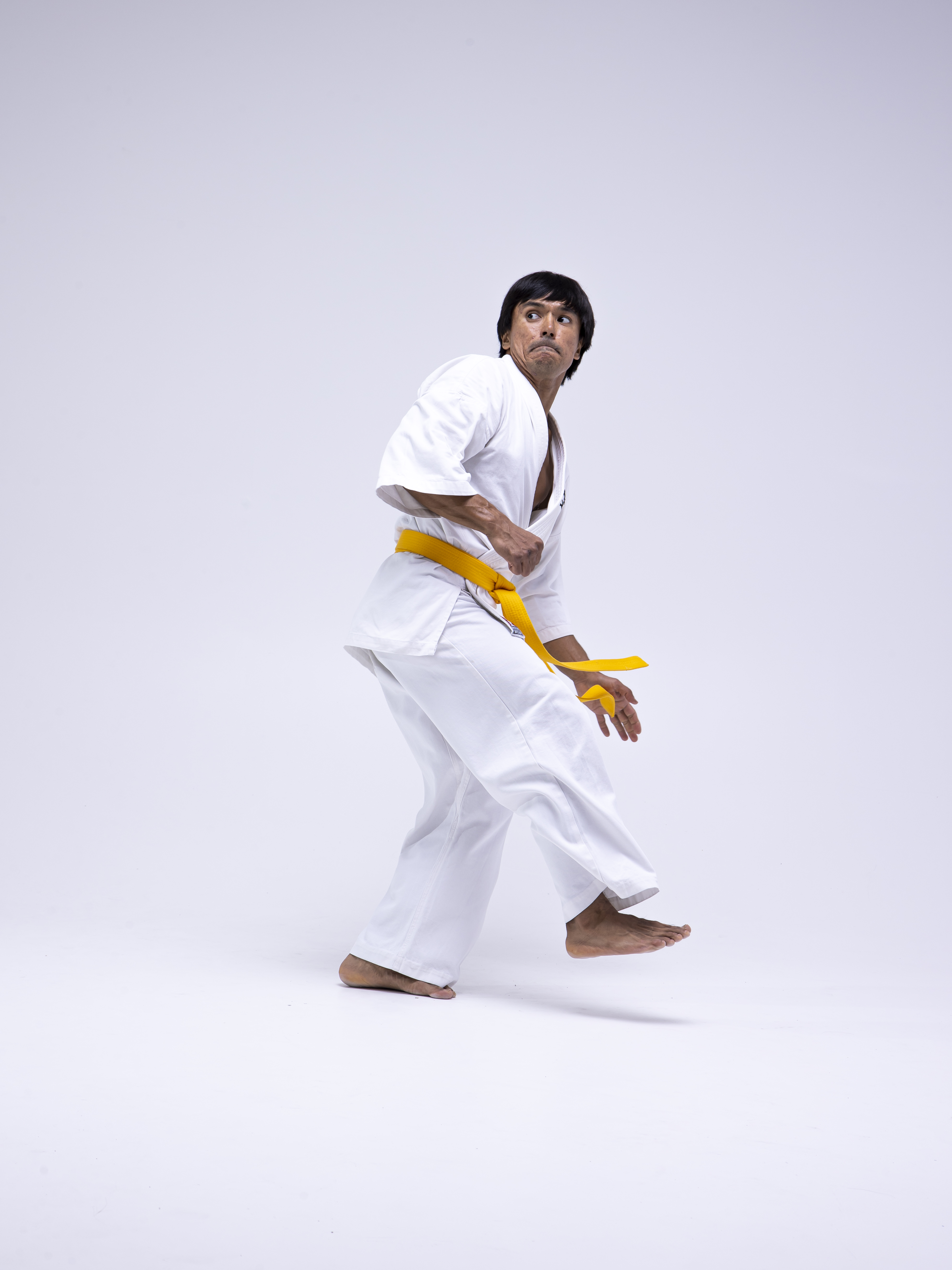 a man wearing a white karate gi with a yellow belt is captured in a martial arts stance. he is barefoot and appears to be kicking, with one foot raised off the ground. the man is looking upwards with an intense expression. the image is a studio shot against a plain white background.