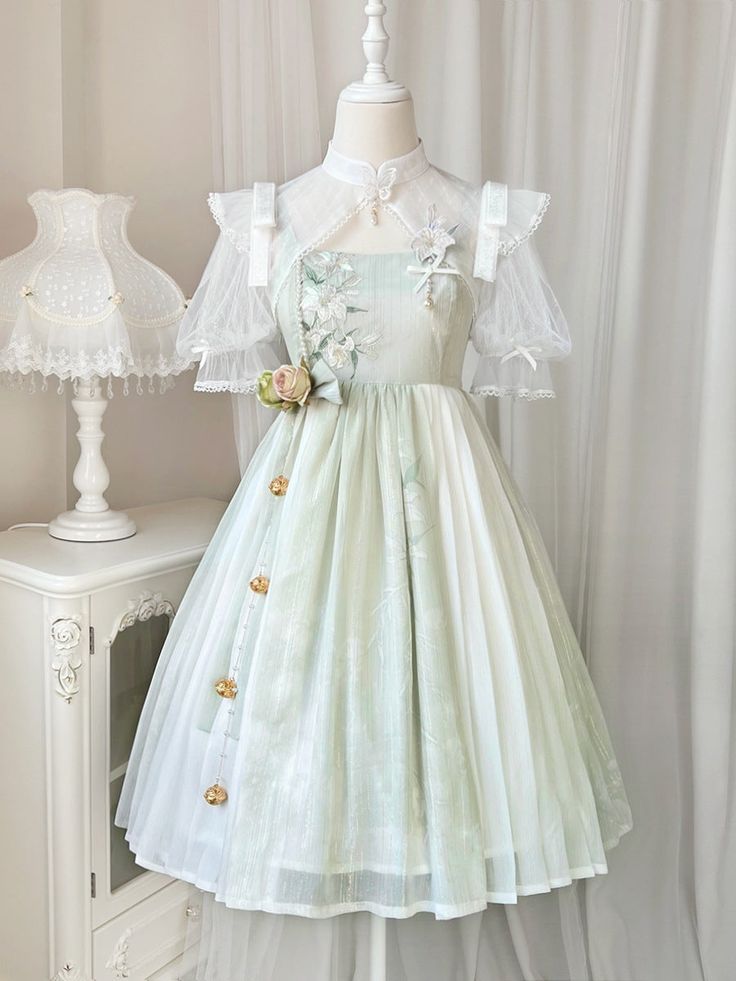 the image shows a light green and white lolita fashion dress displayed on a mannequin. the dress has a floral design and lace accents. the background is a white room with a white vintage nightstand and lamp.