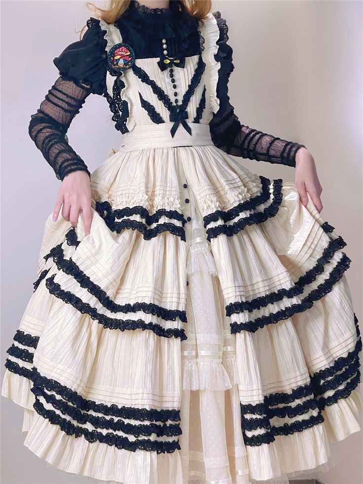 a woman is wearing an elegant, offwhite lolitastyle dress with multiple layers of ruffles and black lace trim. the dress has a fitted bodice and a full, flowing skirt. she is also wearing a black blouse with sheer sleeves underneath.  she is posing with her hands holding out the sides of her skirt. the background is a plain white wall.