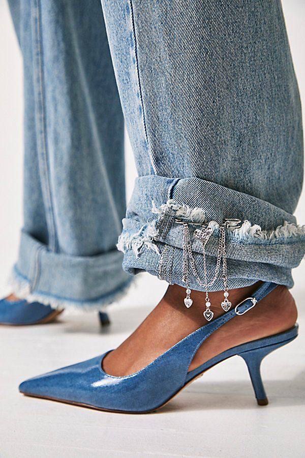 a closeup shot shows a person wearing light wash denim jeans and blue high heels. the jeans are cuffed at the bottom, revealing a raw hem with frayed edges. three silver chains are attached to the cuff, adorned with heart charms. the blue high heels feature a pointed toe and an ankle strap. the background is plain and lightcolored, emphasizing the clothing and footwear.