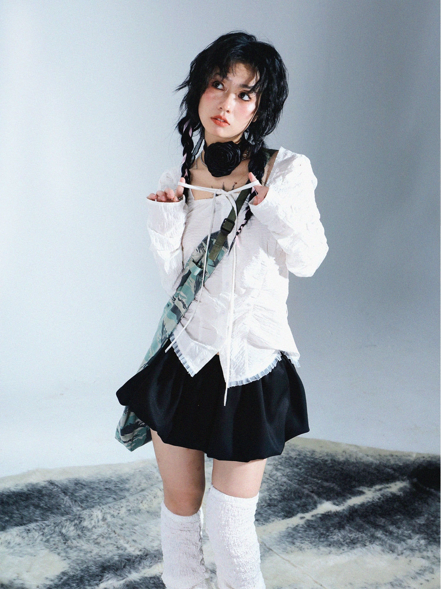 young asian woman with dark hair styled in a bob is posing in a studio setting. she is wearing a white ruffled blouse, a black pleated mini skirt, white kneehigh socks, and a camouflagepatterned shoulder bag.  she has on a dark choker necklace. the background is a plain white with a textured dark rug underfoot.  the overall aesthetic is a blend of cute and grunge.  the image appears to be a fashion photography shot.