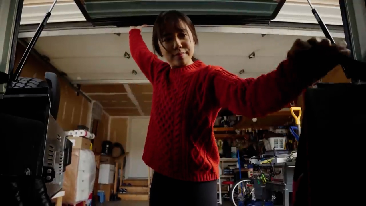 a young woman in a red sweater stands in a garage with the trunk of her car open. she is looking directly at the camera, which is positioned at a low angle, creating a sense of perspective looking up at her. the garage is cluttered with various items, suggesting an activity like packing, unpacking, or organizing. the lighting is natural and diffused, indicating an overcast day.