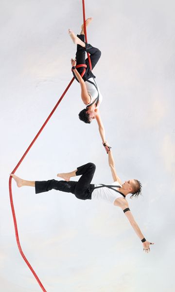 two acrobats are performing a dynamic aerial dance routine, suspended by red ropes. the woman in the white top and black pants is hanging upside down, her leg is hooked around the rope, while the man in the black shirt and black pants, is swinging through the air holding the rope. the photo captures the dynamic and agile movements of the performers as they gracefully navigate their aerial dance routine. the image is shot against a white background that highlights their movements.