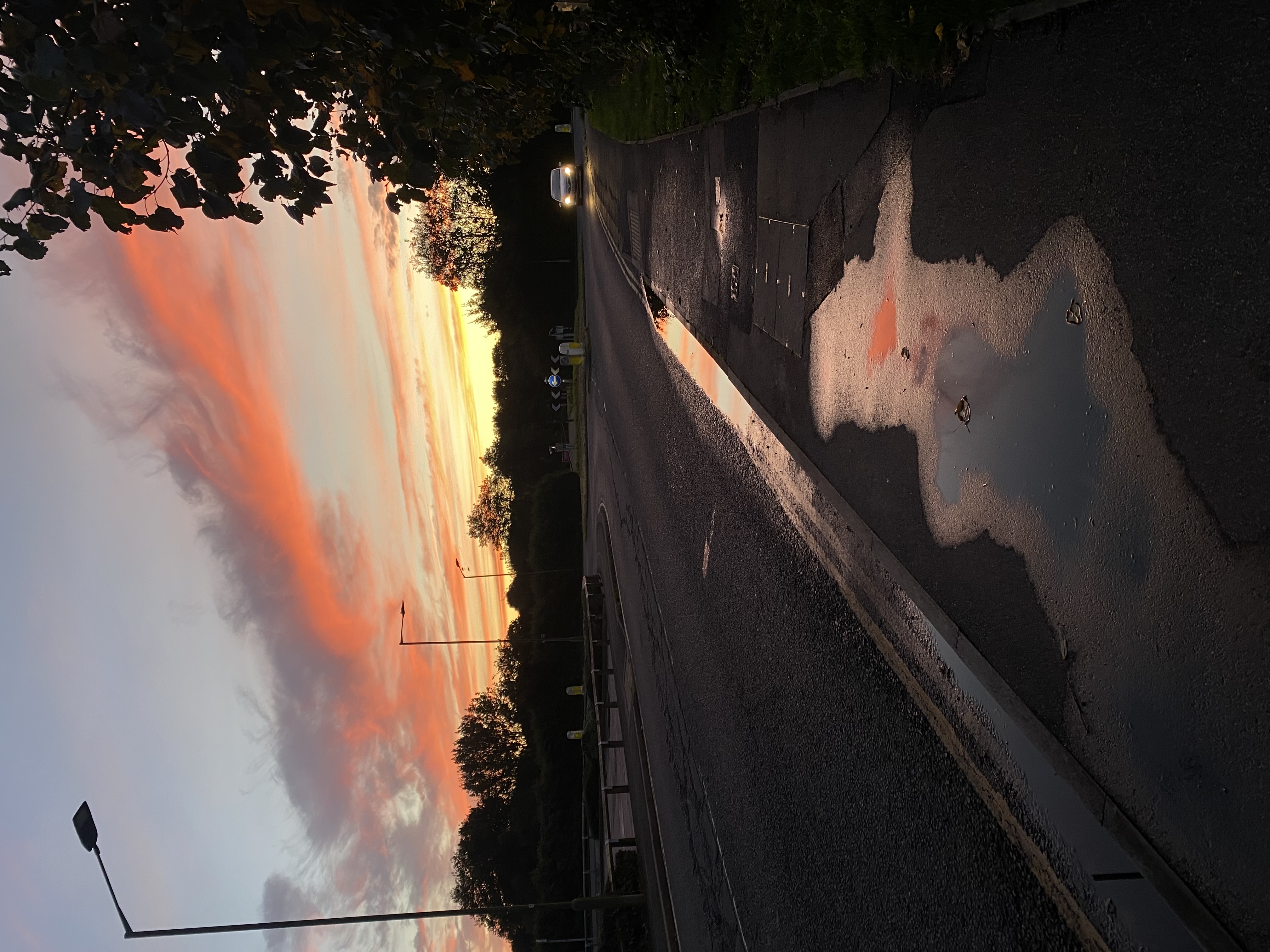 a photo of a road at sunset. the sky is filled with pink and orange clouds, and the sun is setting in the distance. there is a large puddle in the middle of the road, reflecting the sunset colors. there are trees on either side of the road, and street lights are visible in the distance. a single car can be seen driving in the distance.