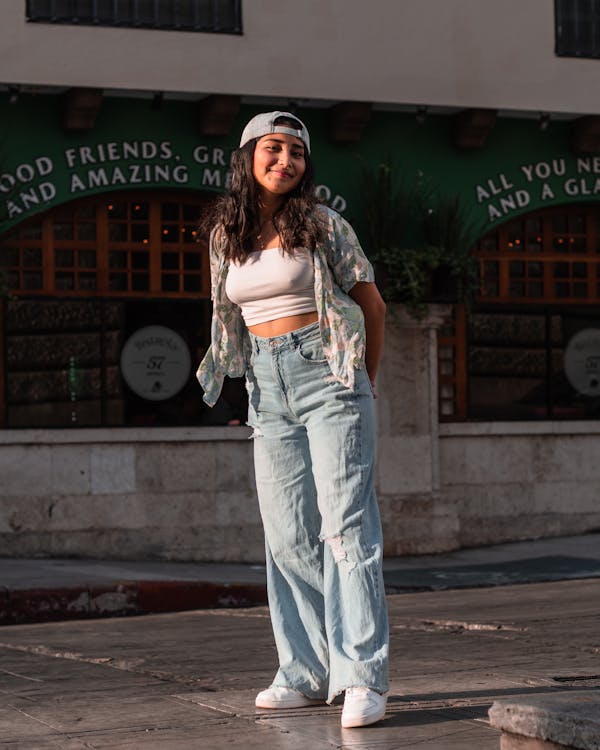 a young woman stands on a city street, smiling at the camera. she is wearing a casual outfit: a white crop top, a floral print shirt, wideleg light blue distressed jeans, a light blue baseball cap, and white sneakers. she is standing in front of a building with a green sign above the entrance. the sun is shining, creating shadows on the ground.