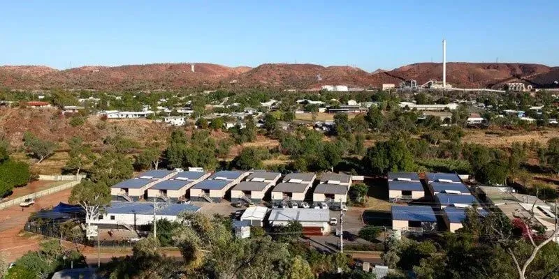 an aerial view of a town in the australian outback. the town is surrounded by red dirt and sparse vegetation. in the distance, there is a large mining operation. there are a number of residential buildings in the town, as well as some commercial buildings. the town appears to be relatively remote and sparsely populated.  the image is taken from a high angle, giving the viewer a panoramic perspective of the town and its surroundings.