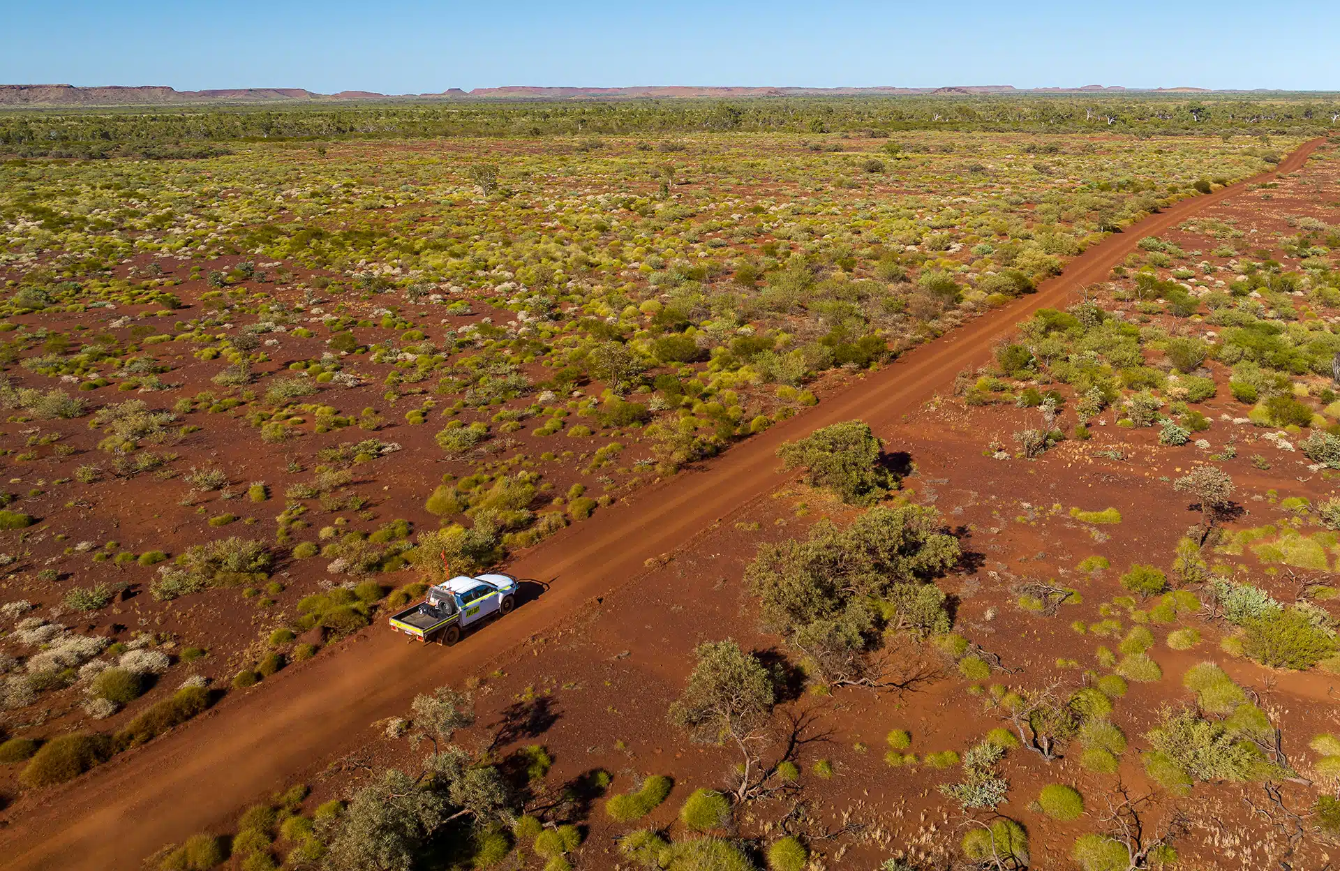 an aerial view of a dirt road winding through the australian outback. the road is surrounded by red dirt and sparse vegetation, creating a desolate and rugged landscape. a 4wd vehicle travels along the road, highlighting the vastness and remoteness of the area. the blue sky above emphasizes the clear atmosphere and the vastness of the desert. the scene evokes a sense of adventure and exploration, inviting viewers to imagine the challenges and beauty of traversing this remote region.