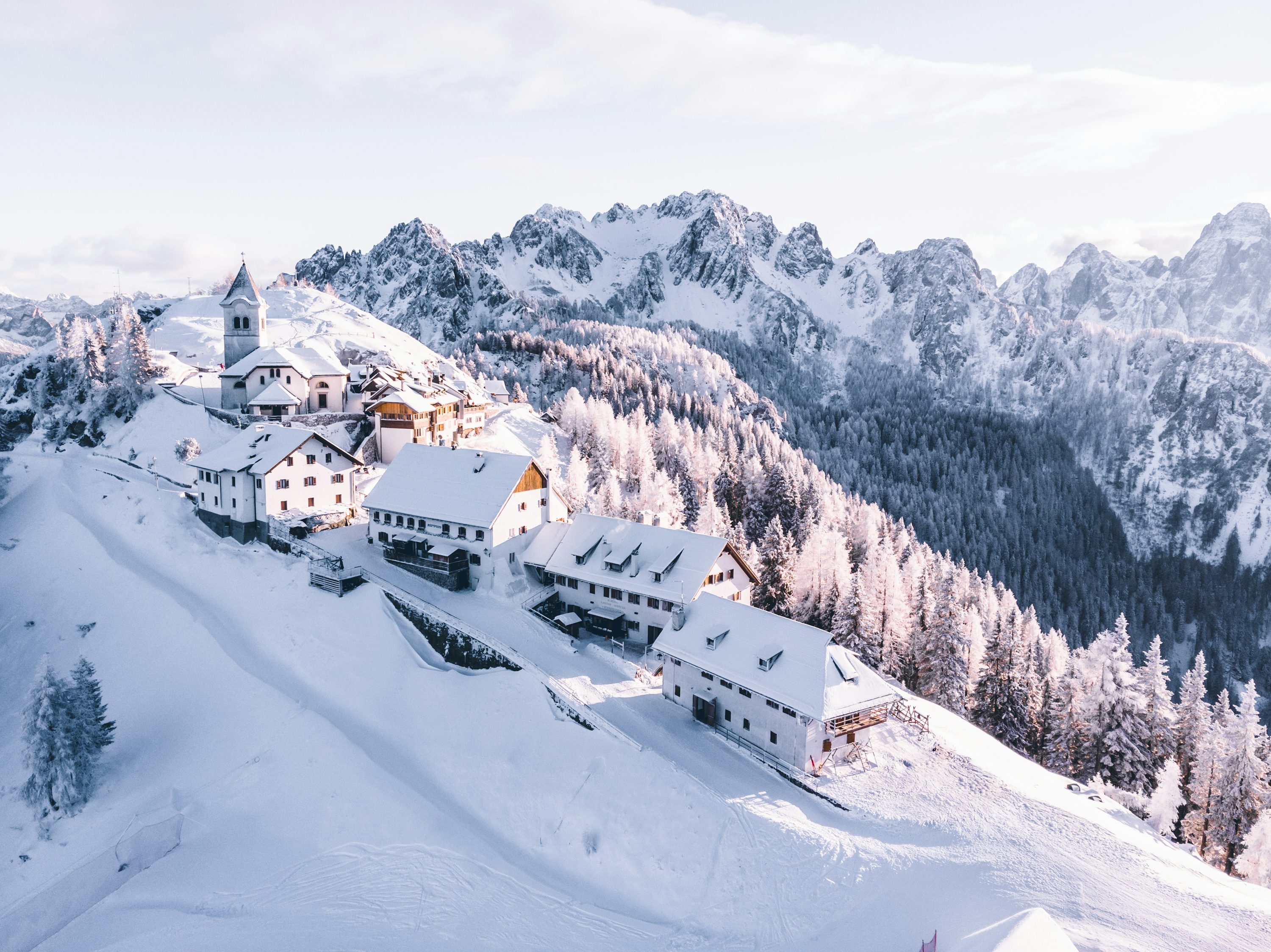 a panoramic view of a snowcovered mountain village nestled in the heart of the alps.  the village is made up of a few white buildings, including a small church, with a few houses clustered around it. the snow is pristine and white, and the trees are covered in a thick blanket of snow. the mountains are towering in the background, and the sky is a clear blue. the scene is serene and peaceful, and the beauty of the winter landscape is evident.  the air is crisp and cold, and the sun is shining brightly on the snow.