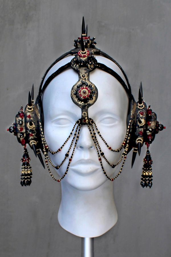 this is a closeup photo of a headpiece made of black, red, and gold materials. the headpiece is adorned with spikes, beads, and decorative elements, giving it a dramatic and intricate design. the headpiece is worn by a mannequin, showcasing its design and detailing.