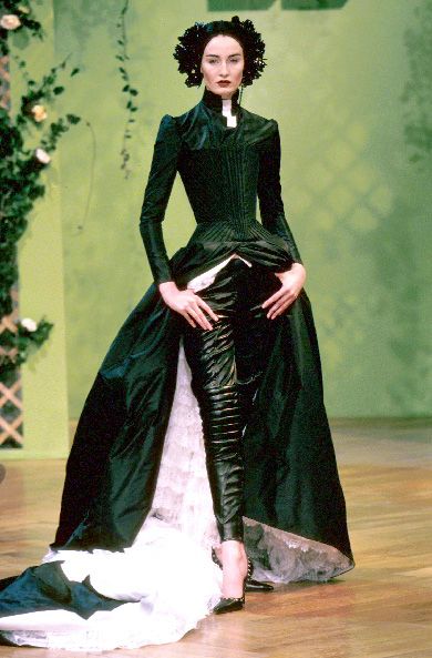 a model is walking the runway wearing a black couture dress with a corset bodice and a long train. she is also wearing black leather pants and high heels. her hair is styled in a headpiece. the background is green.