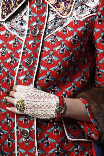 loseup detail shot of a woman's arm and hand wearing a gucci jacket and accessories. the jacket is red with an allover embroidered bee pattern. the gloves are white lace, and the watch is a silver gucci watch with a black band. the background is blurred, and the focus is on the clothing details. the image showcases the rich textures and intricate embroidery of the gucci garments.